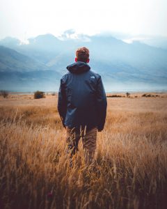 A young man wearing a jacket from The North Face stands in a field of brown grass and looks out at the mountains beyond.