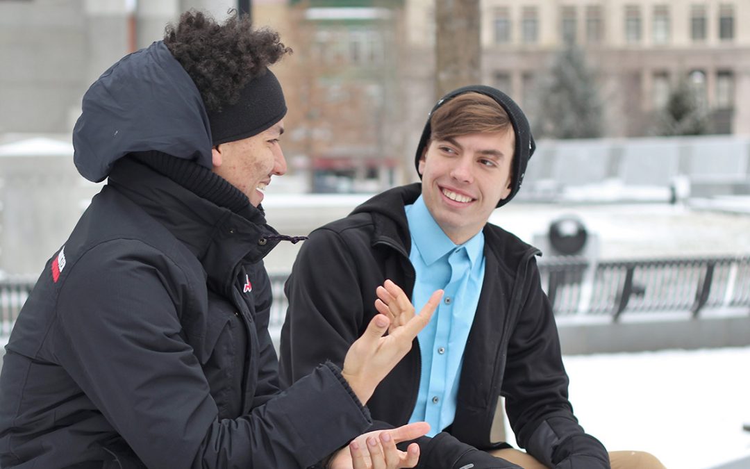 Two young, male college students talk on a bench at their school in the snow.