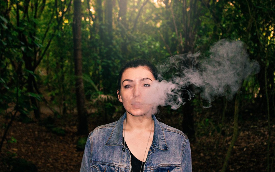 Young woman blowing e-cigarette vapor in front of trees.