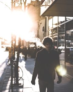 Young man blows vapor from an e-cigarette as he walks down the sidewalk at sunset.